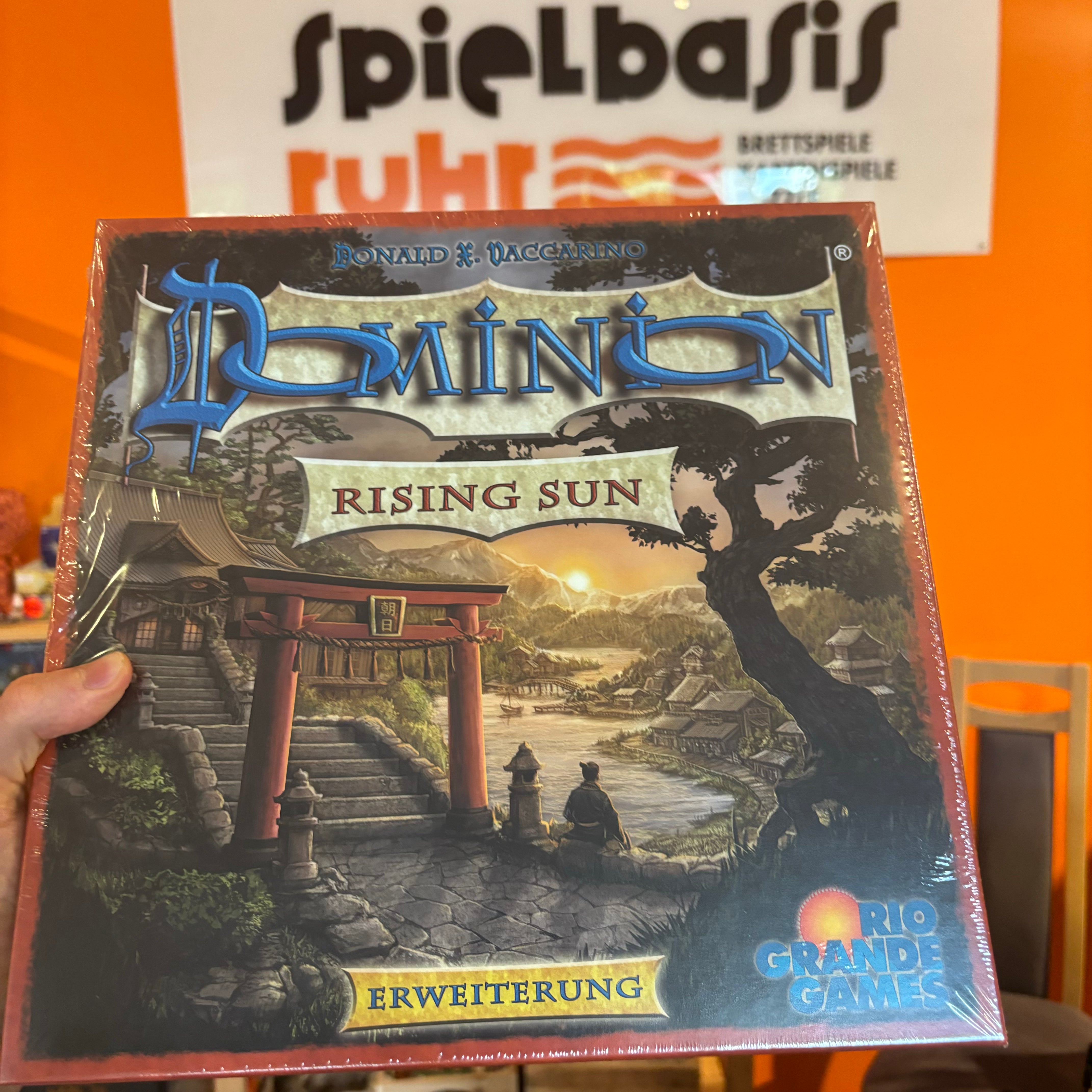 Dominion - Rising Sun Erweiterung Cover der Spielschachtel; Haus bei Sonnenuntergang vor Dorf und Bergen im asiatischen Setting